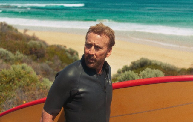 Nicolas Cage as The Surfer in The Surfer. He is wearing a black wetsuit and carrying a yellow surfboard