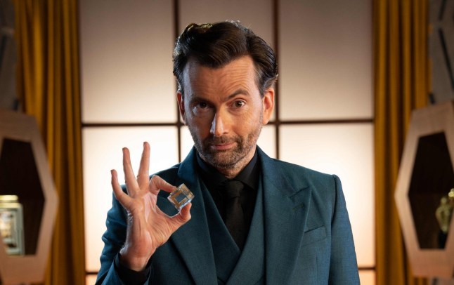 David Tennant in a blue suit smiles at the camera while holding a glass cube in his fingers in promotional imagery for Genius Game